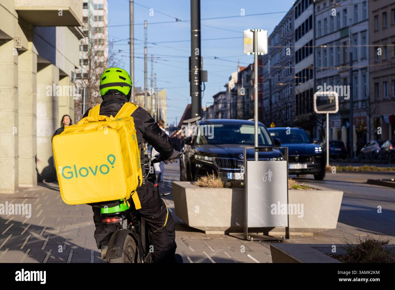 Poznan, Polonia - 8 aprile 2025: Corriere alimentare per Glovo che indossa un casco verde e un vestito nero con un grande zaino giallo viaggia attraverso una U Foto Stock