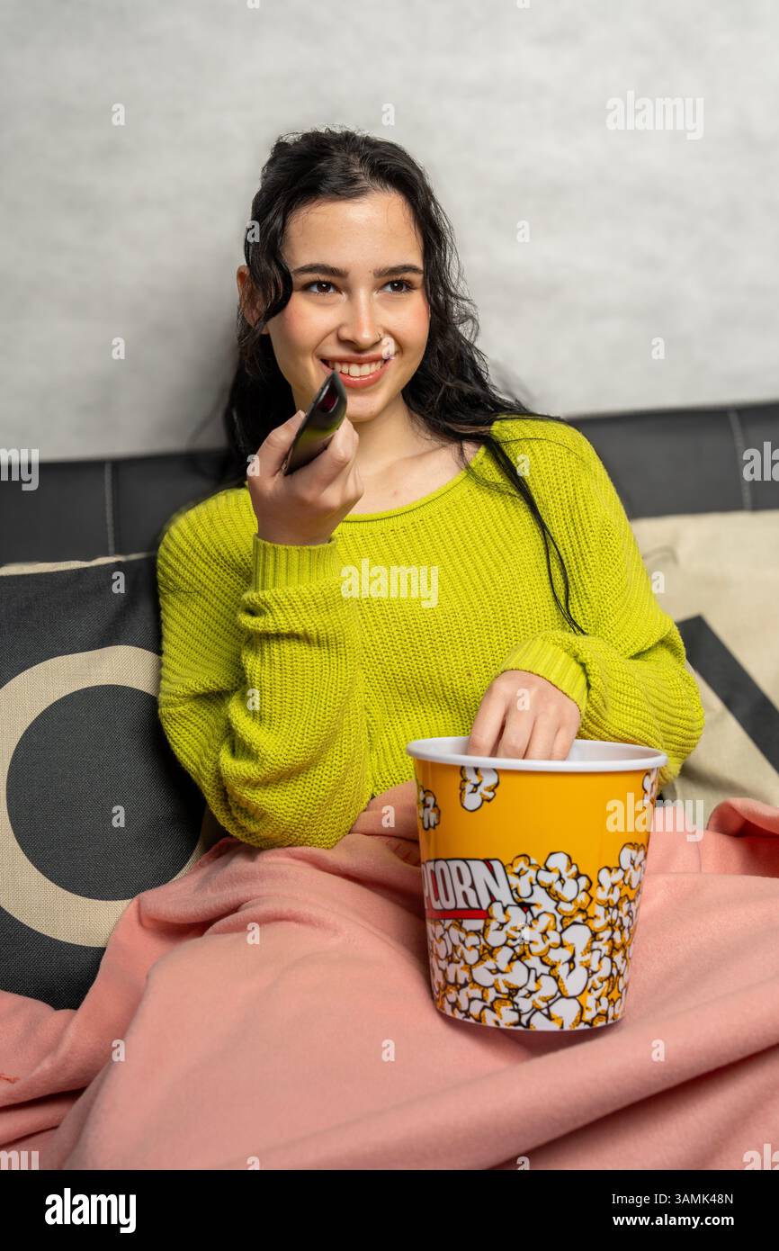 Donna sorridente che usa il telecomando e mangia popcorn mentre guarda la televisione Foto Stock