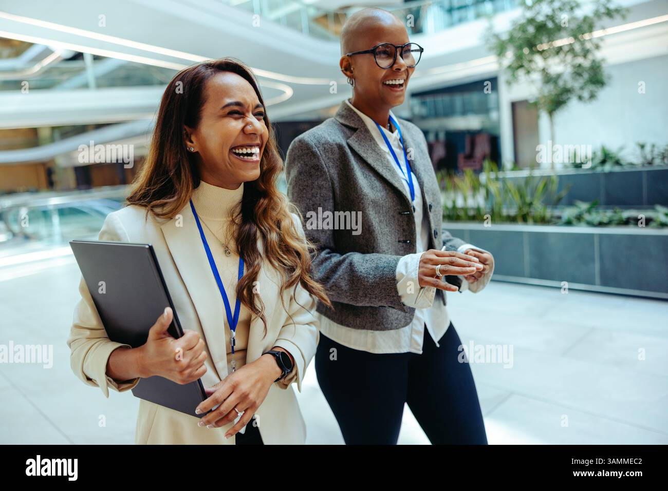 Due allegre donne professionisti che camminano all'interno in un ambiente aziendale. Sono impegnati nella conversazione, indossando sia abiti da lavoro che lanya Foto Stock