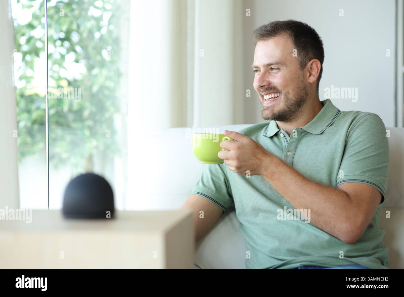 Uomo felice che beve un caffè rilassante con un altoparlante intelligente seduto sul divano di casa Foto Stock