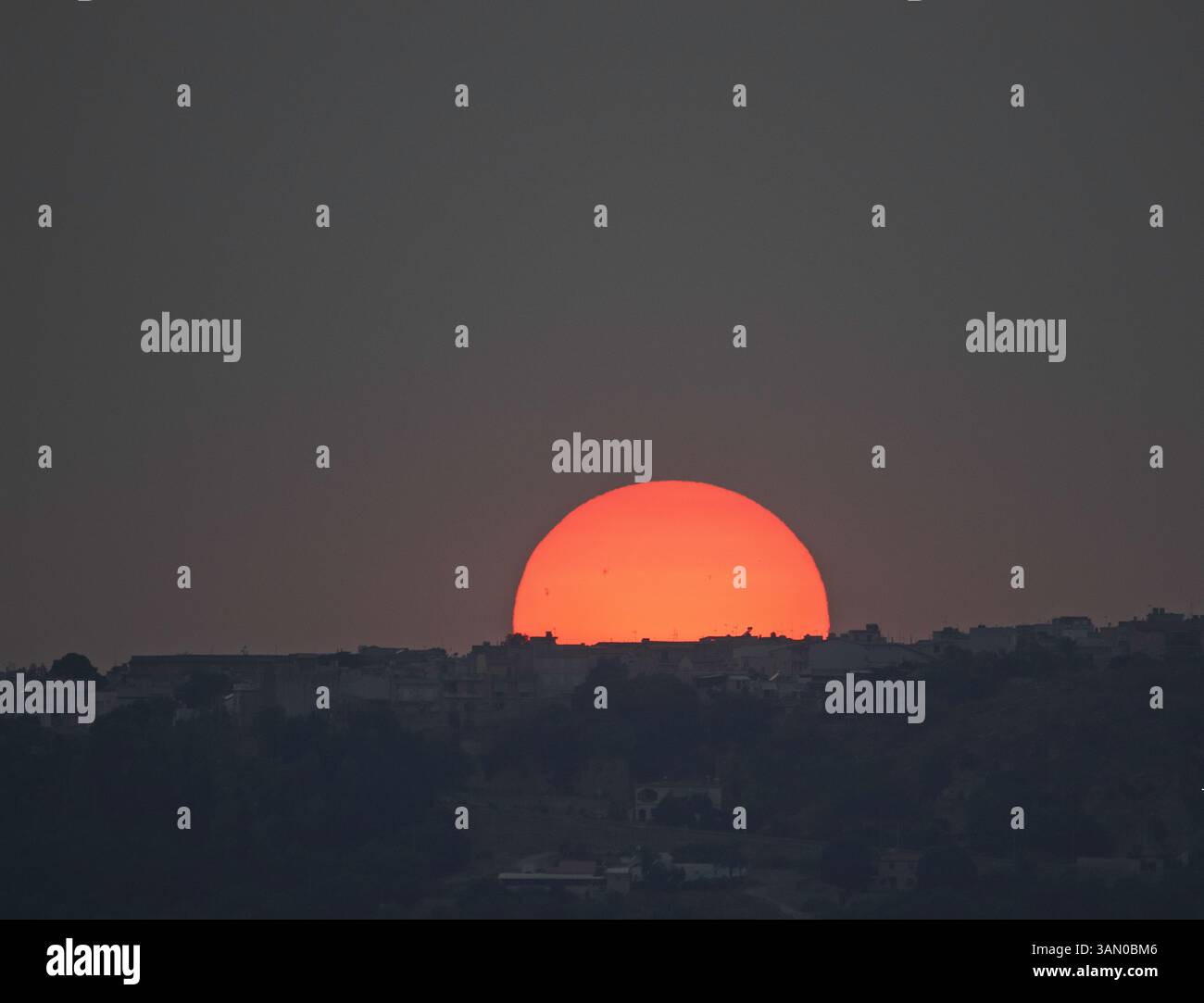 "Il sole tramonta dietro un villaggio collinare in Sicilia, Italia, creando una silhouette serena con morbide tonalità arancioni. Un tranquillo paesaggio mediterraneo al crepuscolo. Foto Stock