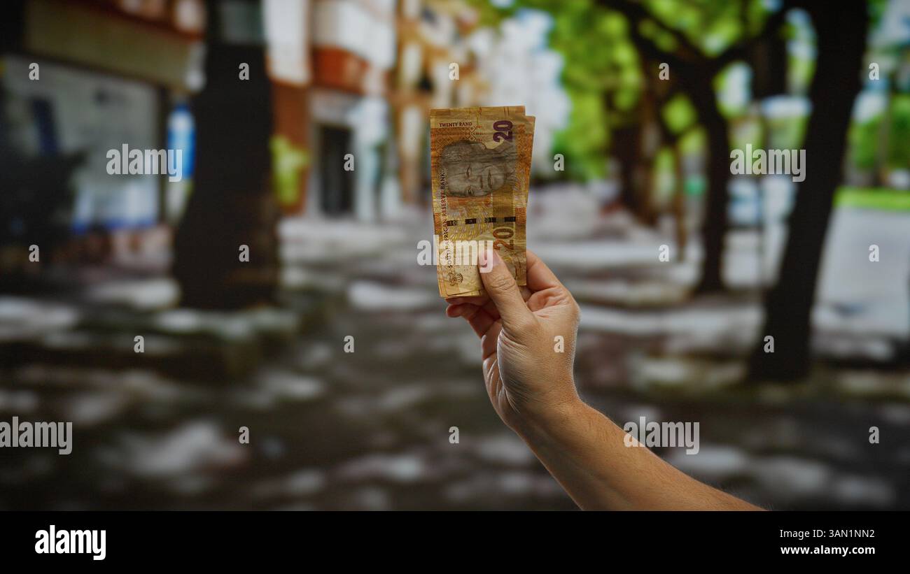 Mano che tiene in mano banconote in rand sudafricane sullo sfondo urbano di strade all'aperto, simboleggiando le transazioni finanziarie o il cambio valuta in un bustlin Foto Stock