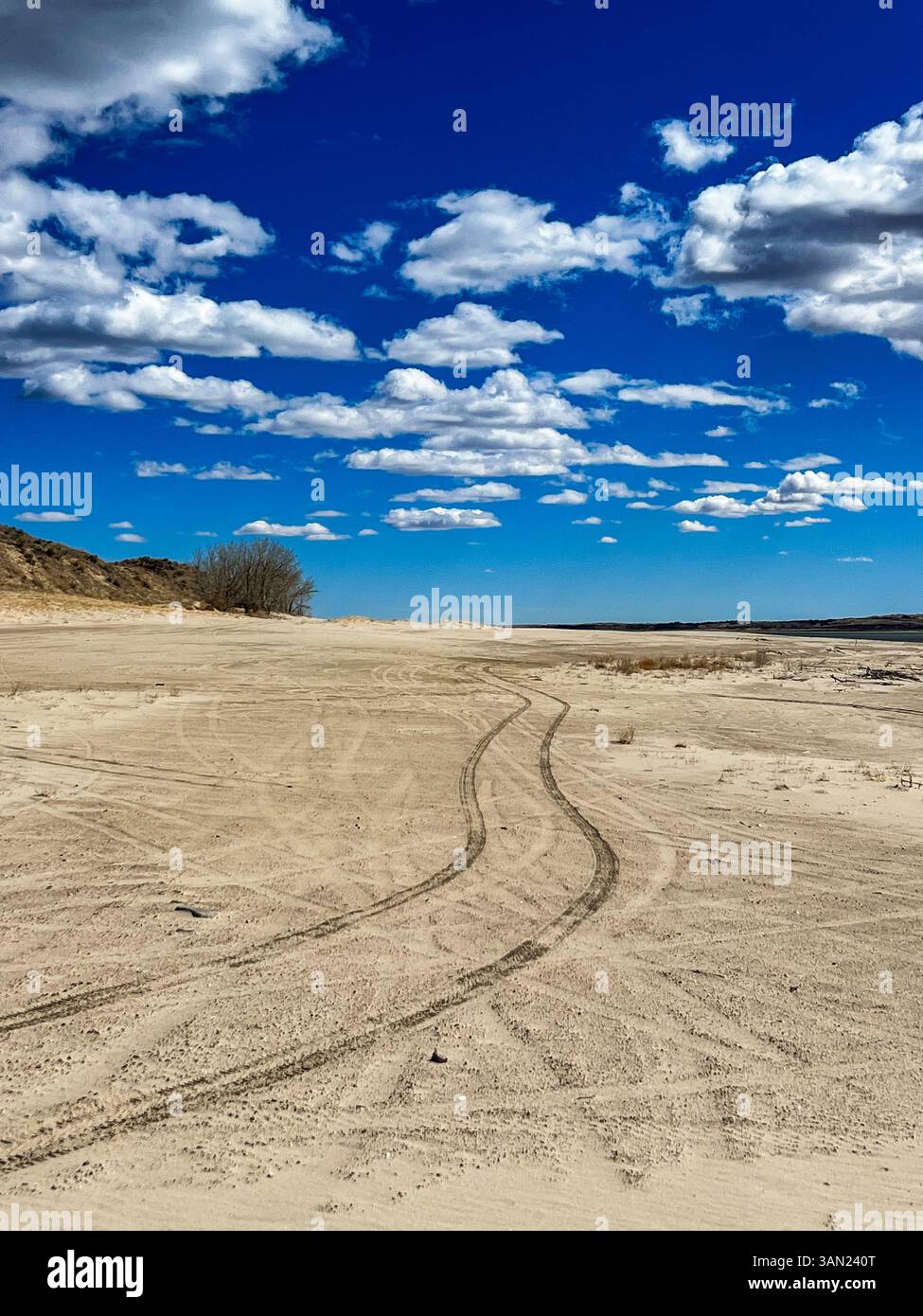 I segni degli pneumatici si curano attraverso la sabbia soffice mentre le nuvole sparse si spostano attraverso un cielo blu brillante, catturando la tranquilla bellezza del paesaggio aperto. - Immagine stock catturata con smartphone