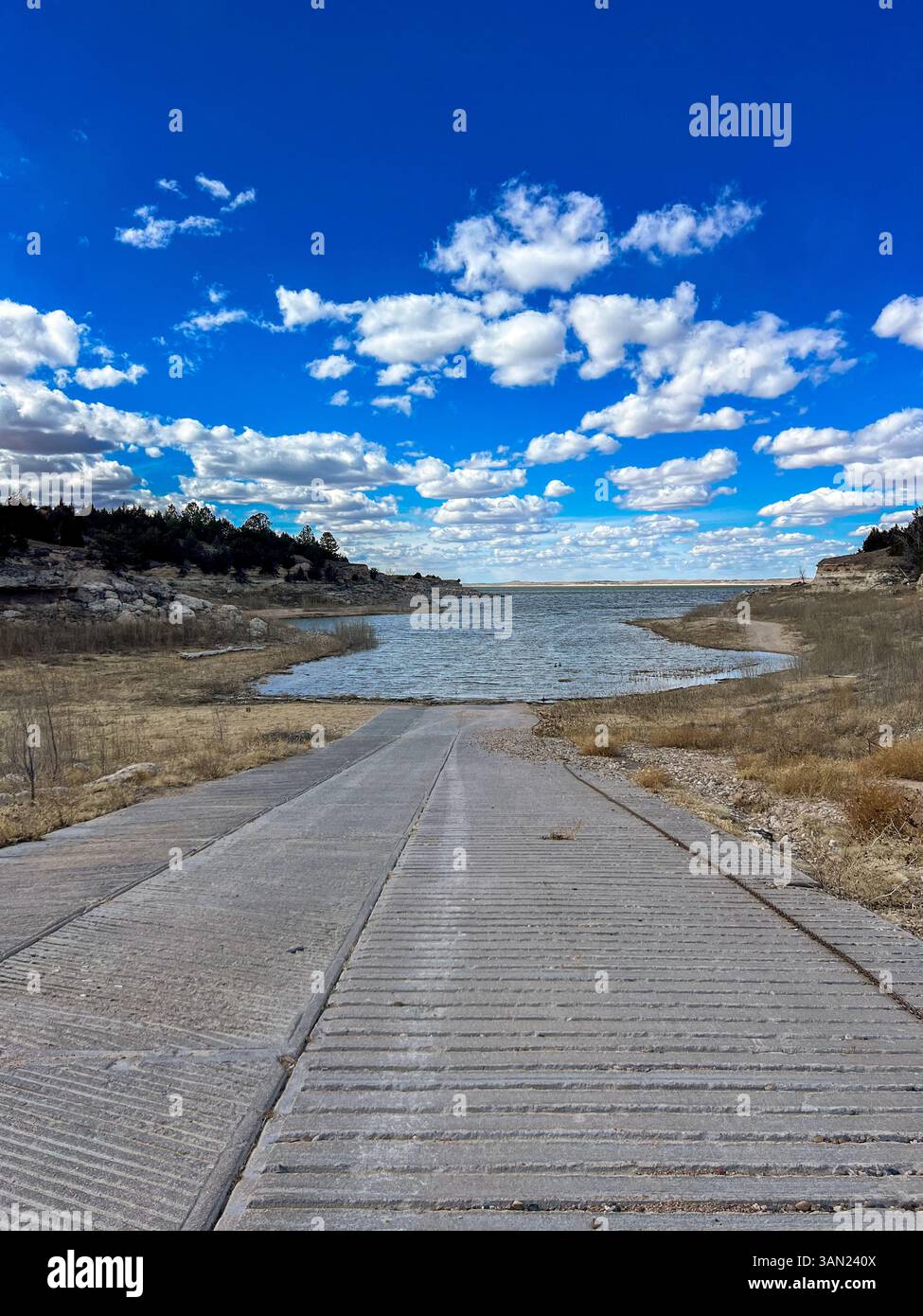Una rampa per imbarcazioni in cemento si estende verso un lago tranquillo, incorniciato da coste rocciose e da un ampio cielo aperto pieno di morbide nuvole alla deriva. - Immagine stock catturata con smartphone