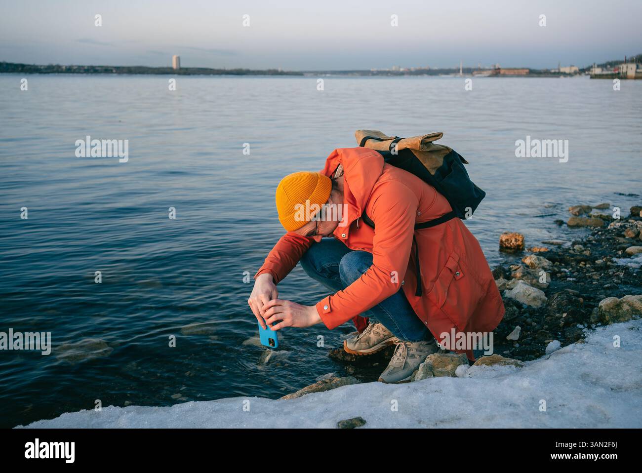 Giovane scienziato accovacciato lungo un fiume in inverno, raccogliendo un campione d'acqua con uno smartphone e una custodia impermeabile, mostrando lavoro sul campo e monitoraggio ambientale Foto Stock