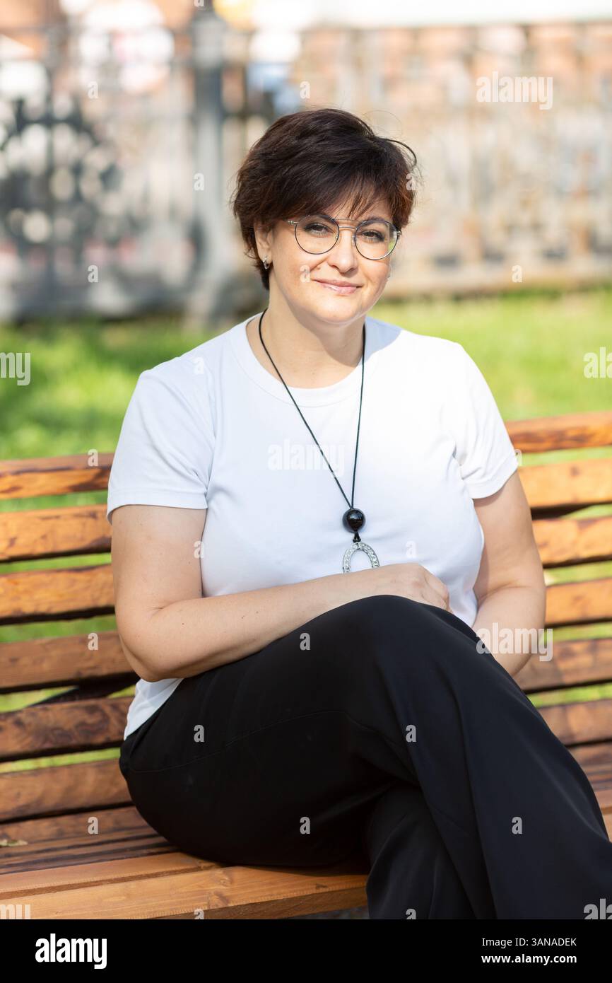 Una bella donna di mezza età con occhiali e capelli scuri corti è seduta su una panchina Foto Stock