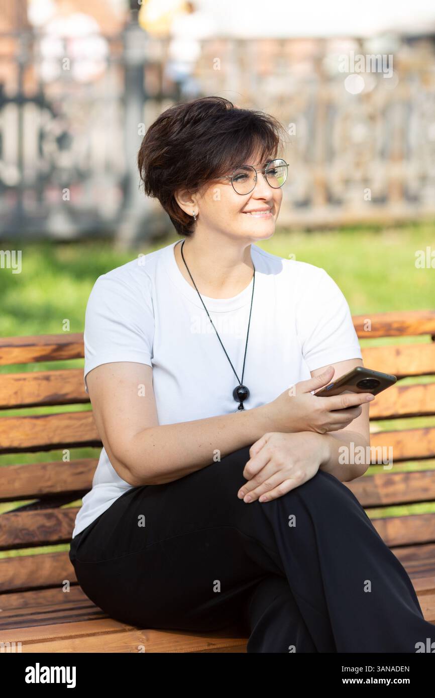 Una bella donna di mezza età con occhiali e capelli scuri corti è seduta su una panchina Foto Stock