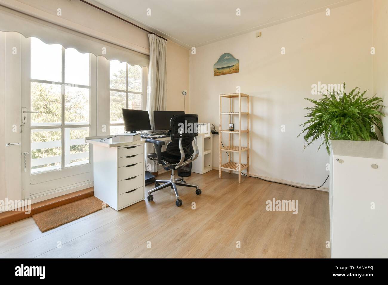 Un elegante home Office con scrivania con due monitor, una comoda sedia e una pianta, il tutto inondato di luce naturale attraverso le grandi finestre. Foto Stock