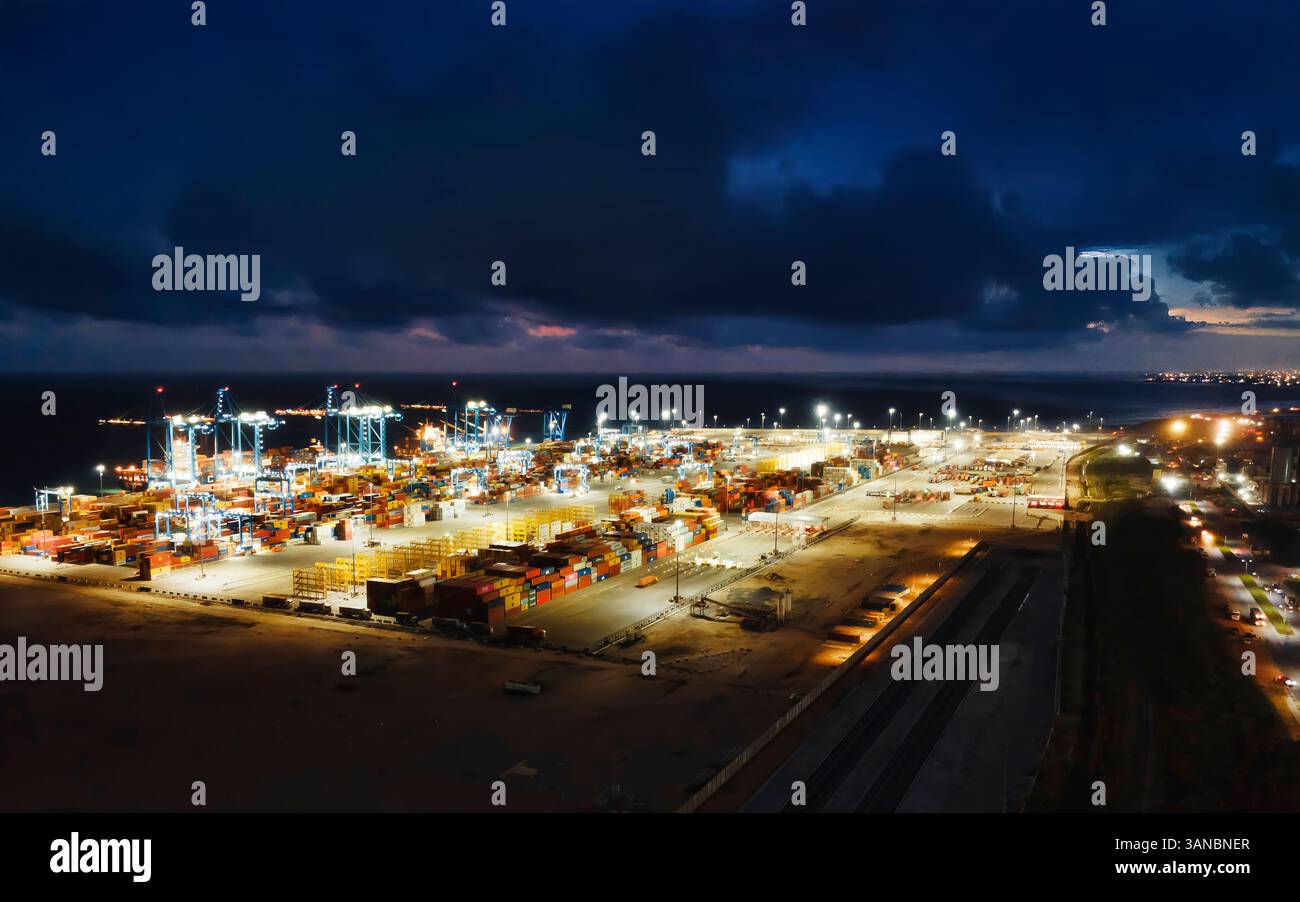 Vista notturna di tema Harbor a tema, Ghana, che mostra l'attività di movimentazione dei container e le operazioni di spedizione sotto un cielo buio e nuvoloso. Le luci accese illuminano l'ampia infrastruttura, catturando la portata e l'importanza di questo importante centro marittimo. Foto Stock