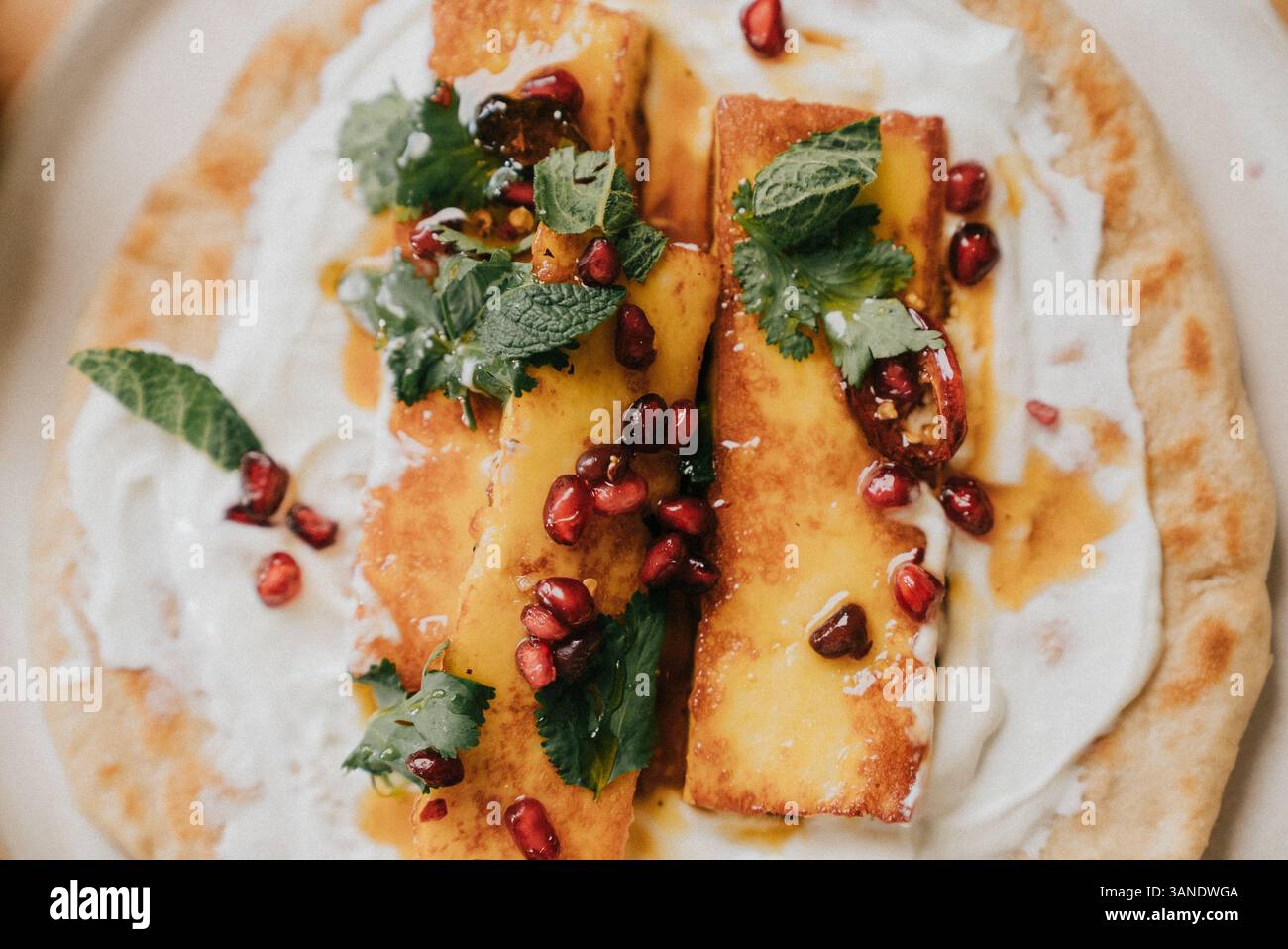 Un piatto di cibo con una base bianca e un contorno di erbe verdi e frutta rossa. Il piatto è una focaccia con una salsa cremosa e un condimento di melograno Foto Stock