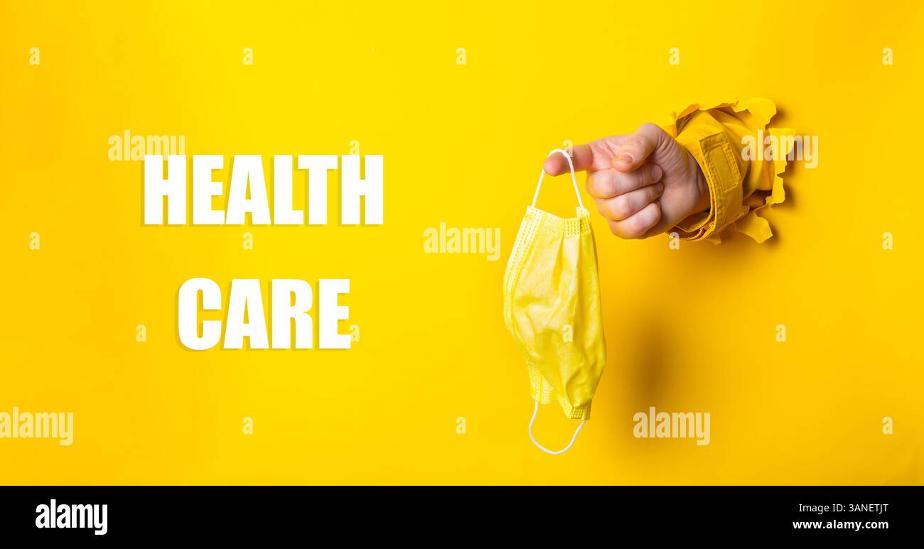Persona che tiene una maschera gialla con la parola assistenza sanitaria scritta di seguito. Concetto di cautela e responsabilità nei confronti della salute pubblica Foto Stock