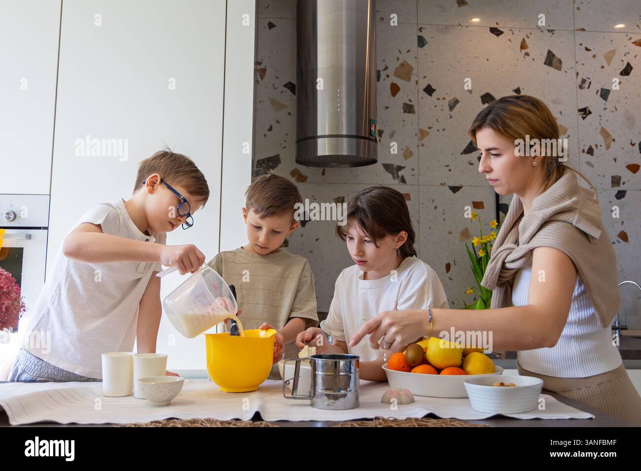 Madre e figli cucinano insieme in cucina. Una madre e i suoi tre figli cuocono insieme in una cucina moderna, mescolando gli ingredienti in ciotole e. Foto Stock