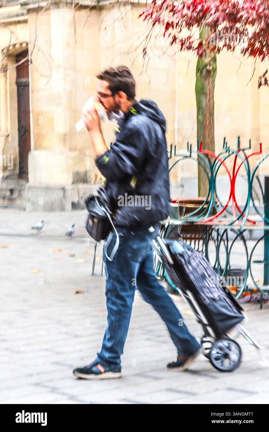 Uomo che mangia panini e tira bagagli a rotelle mentre cammina rapidamente producendo sfocature d'azione - Bruxelles, Belgio. Foto Stock