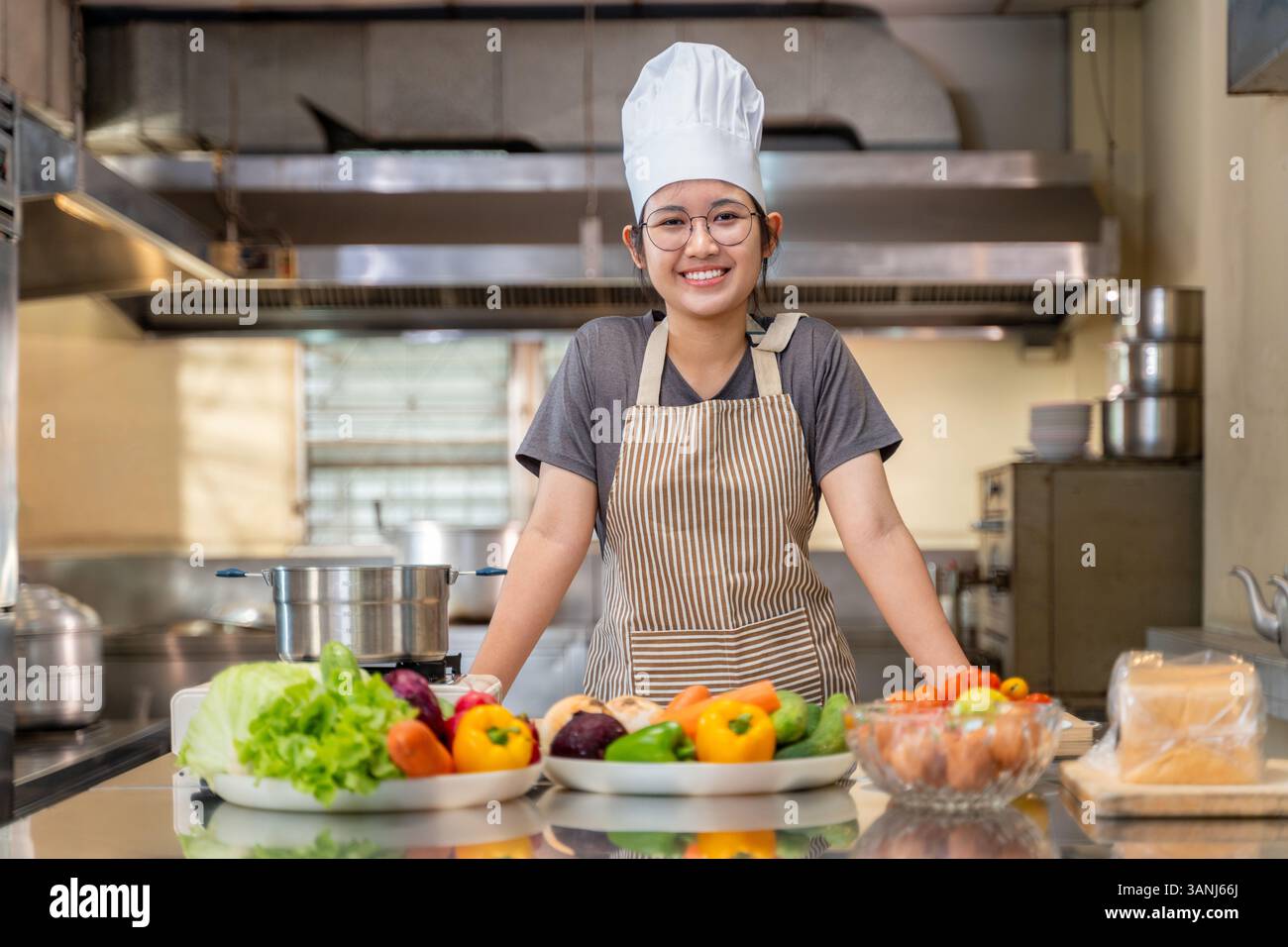 Ritratto di una giovane chef donna in una cucina professionale, in piedi dietro un bancone pieno di verdure fresche e ingredienti da cucina. Indossa una S. Foto Stock