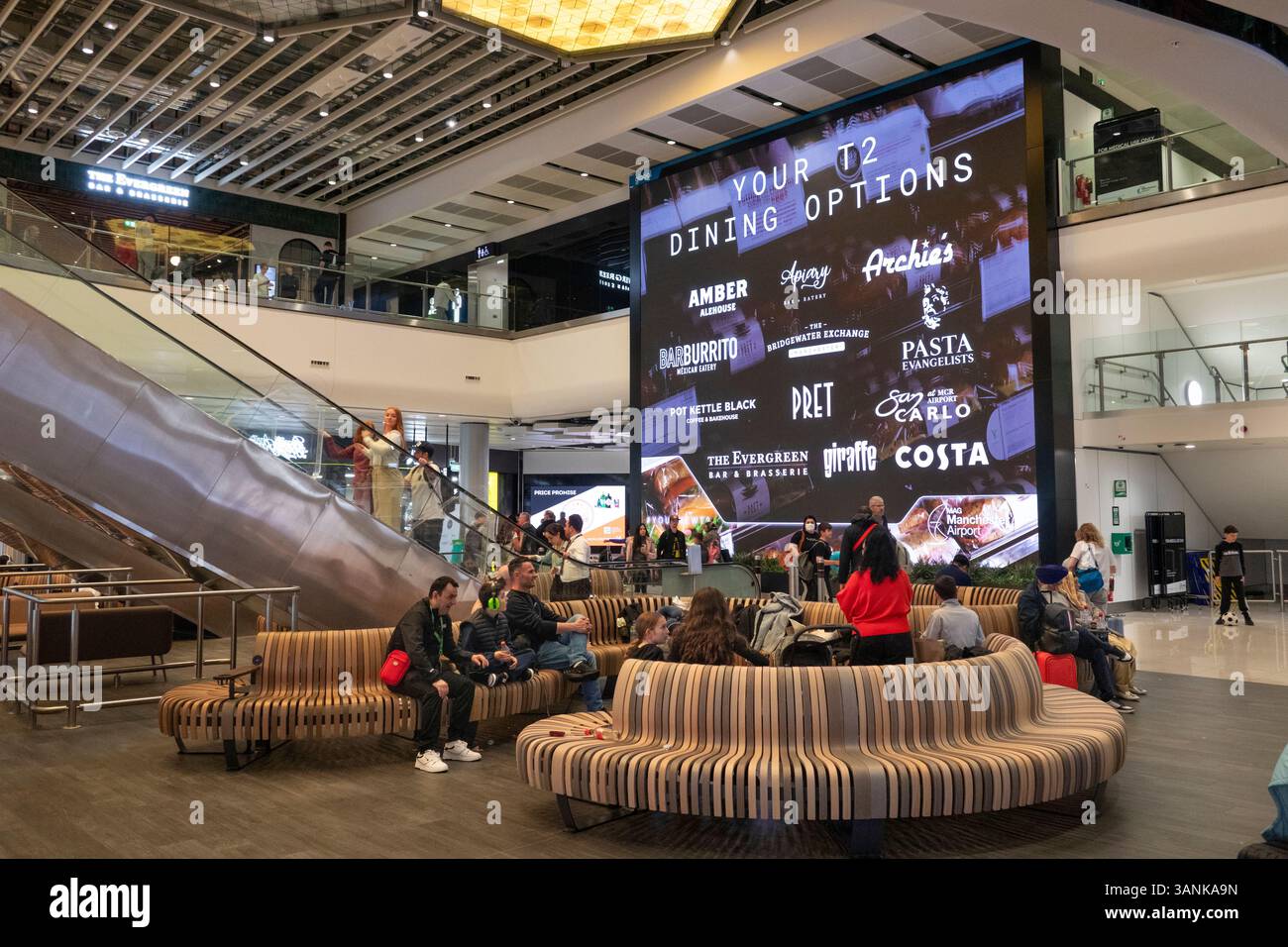 Regno Unito, Inghilterra, aeroporto di Manchester, sala partenze Terminal 2, passeggeri in attesa su un gigantesco schermo pubblicitario Foto Stock
