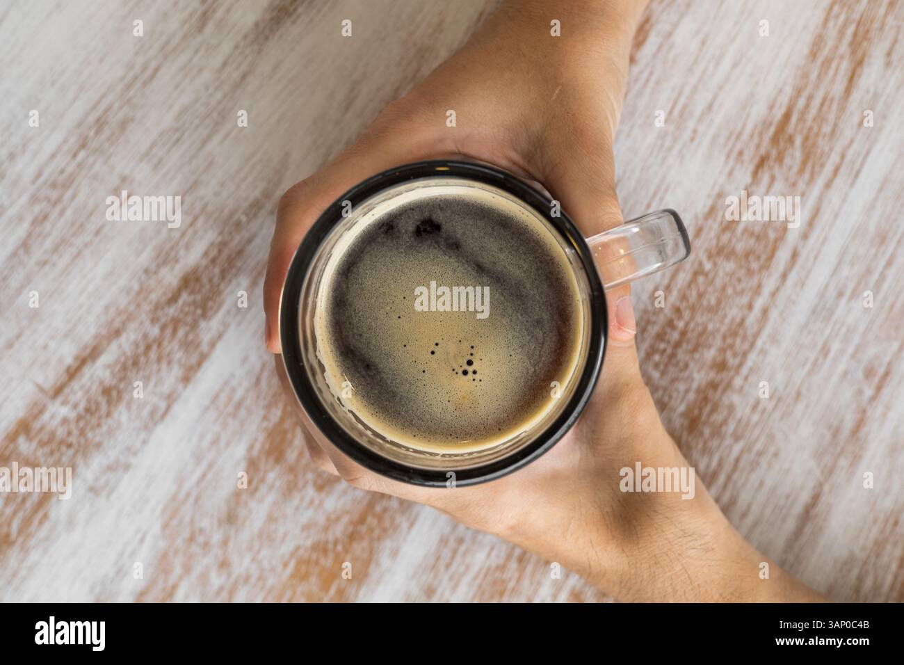 Una persona tiene delicatamente una tazza calda con entrambe le mani su un tavolo di legno con sfumature di crema e miele, che evocano comfort e calma. Foto Stock