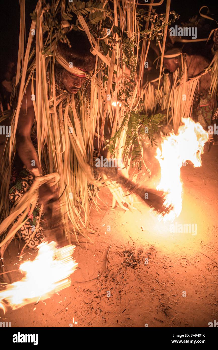 Partecipa a una cerimonia Bwiti, vestito con un costume rituale fatto di foglie di palma essiccate, preparando il fuoco per iniziare la sua danza. Foto Stock