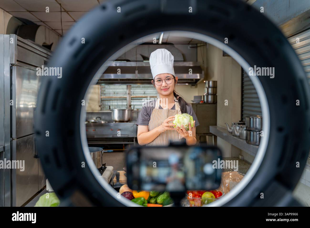 Una giovane chef donna che indossa un grembiule a righe e un cappello da cuoco bianco registra un tutorial di cucina utilizzando uno smartphone e una luce anulare in un gattino commerciale Foto Stock
