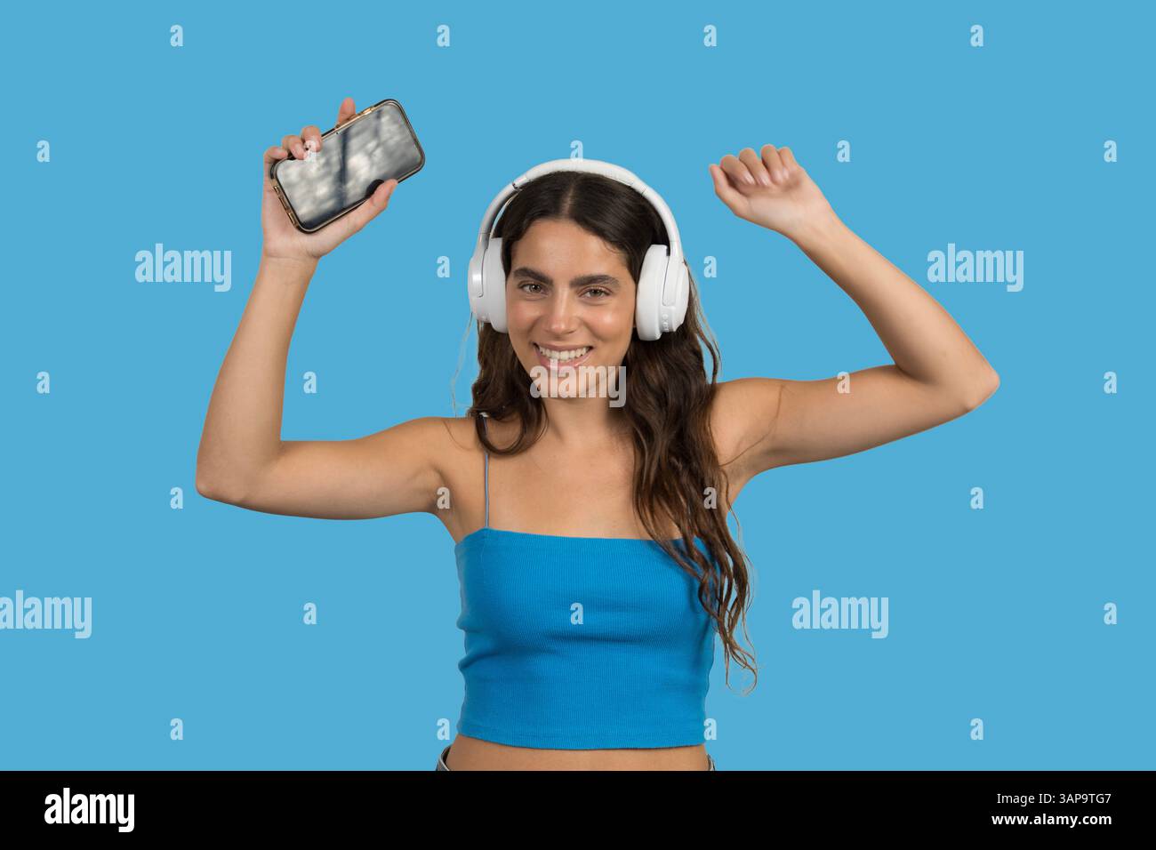 Giovane donna felice che indossa le cuffie per ascoltare musica su sfondo blu, tenere lo smartphone e ballare Foto Stock