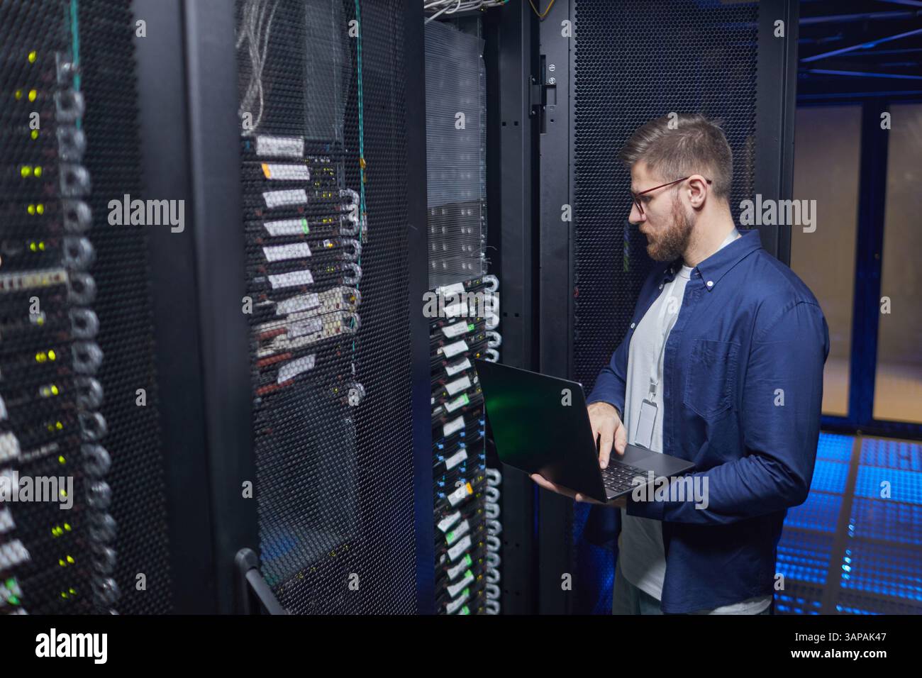 Tecnico in piedi nella sala server per controllare le connessioni e lo stato dei server utilizzando un computer portatile, circondato da più rack di apparecchiature e cavi Foto Stock