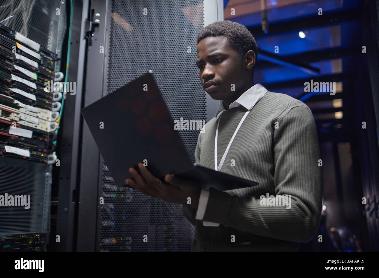 Tecnico afroamericano che lavora con un laptop all'interno della sala server del data center, analizzando le prestazioni del server, monitorando sistemi e infrastrutture, garantendo stabilità e connettività di rete Foto Stock