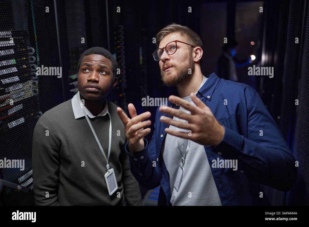 Due tecnici nel data center, uno afroamericano, in piedi vicino ai rack server mentre discutono del processo di manutenzione, una persona che gestiva con la mano, mostrando attenzione Foto Stock