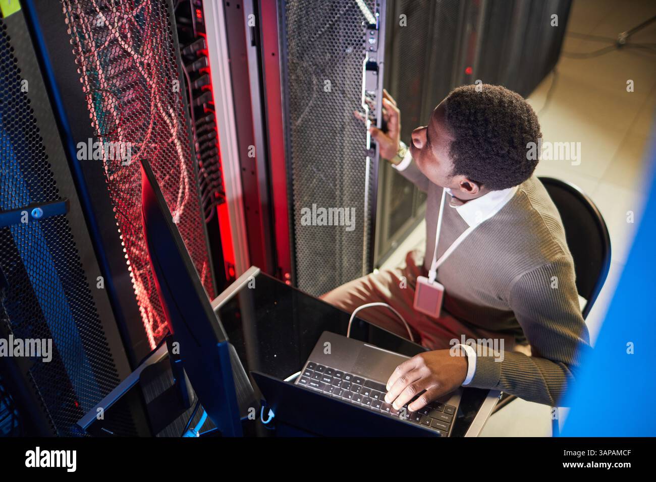 Uomo afroamericano che monitora le apparecchiature della sala server nel data center, seduto con un laptop, garantendo che i sistemi siano operativi e sicuri Foto Stock