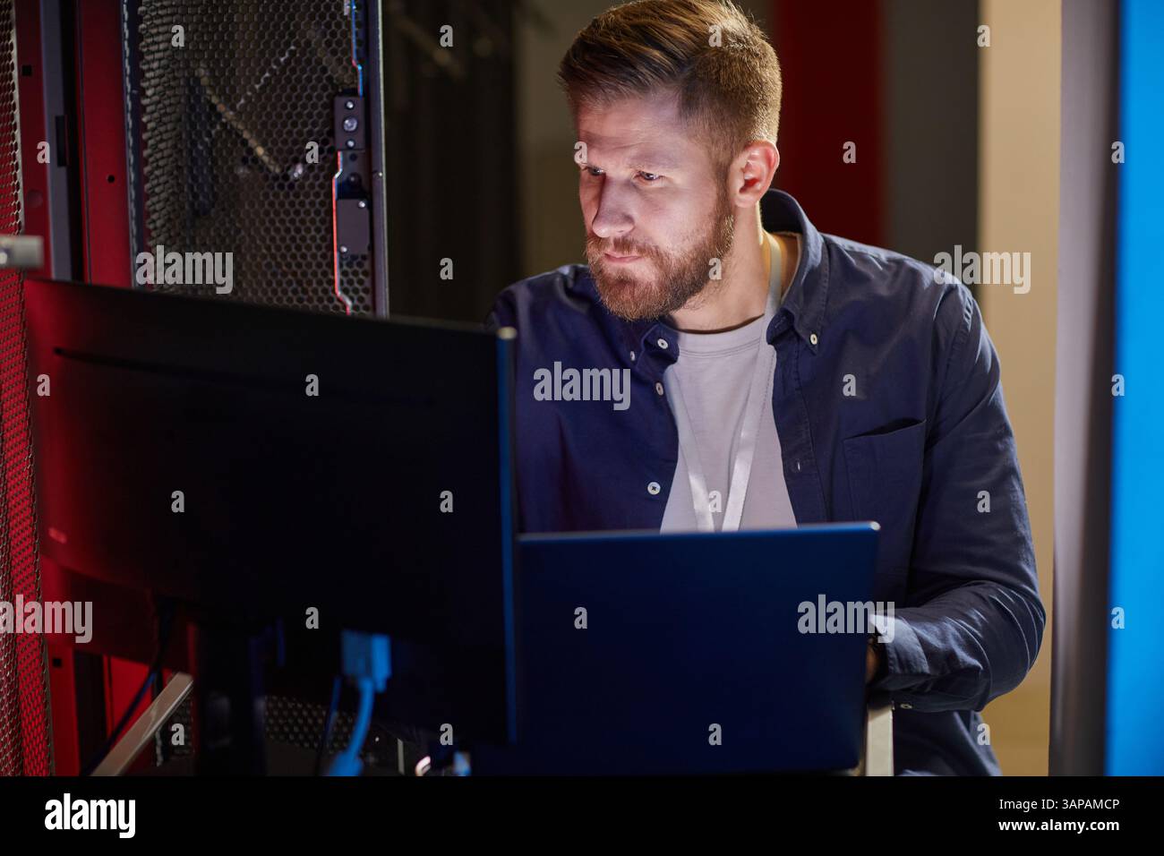 Uomo con barba che osserva lo schermo del computer in sala server con apparecchiature tecnologiche che lo circondano e un'intensa concentrazione sul viso Foto Stock