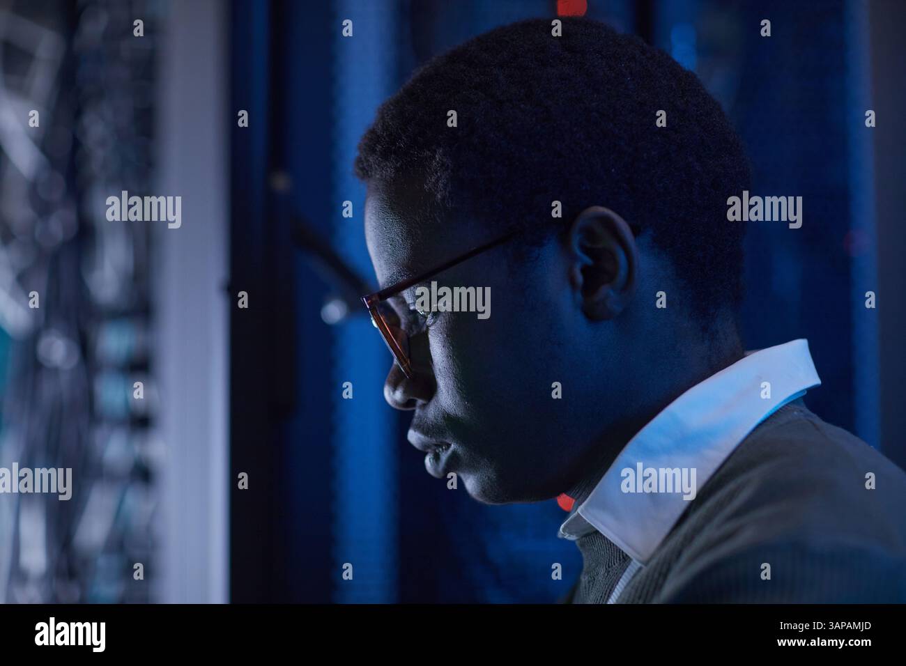 Vista del profilo di un tecnico dedicato che esamina la rete informatica in una sala server in condizioni di scarsa illuminazione. Intensa concentrazione sulle attrezzature tecnologiche Foto Stock