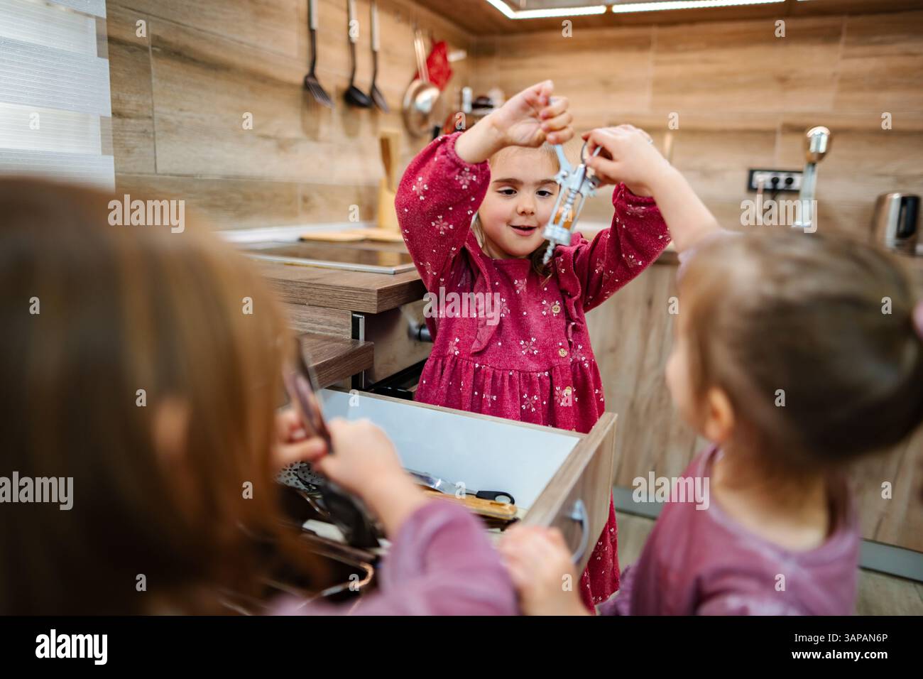 Tre bambini si riuniscono intorno a un cassetto della cucina, pieno di vari utensili. Un bambino tiene in mano un misurino, condividendo il divertimento della scoperta Foto Stock