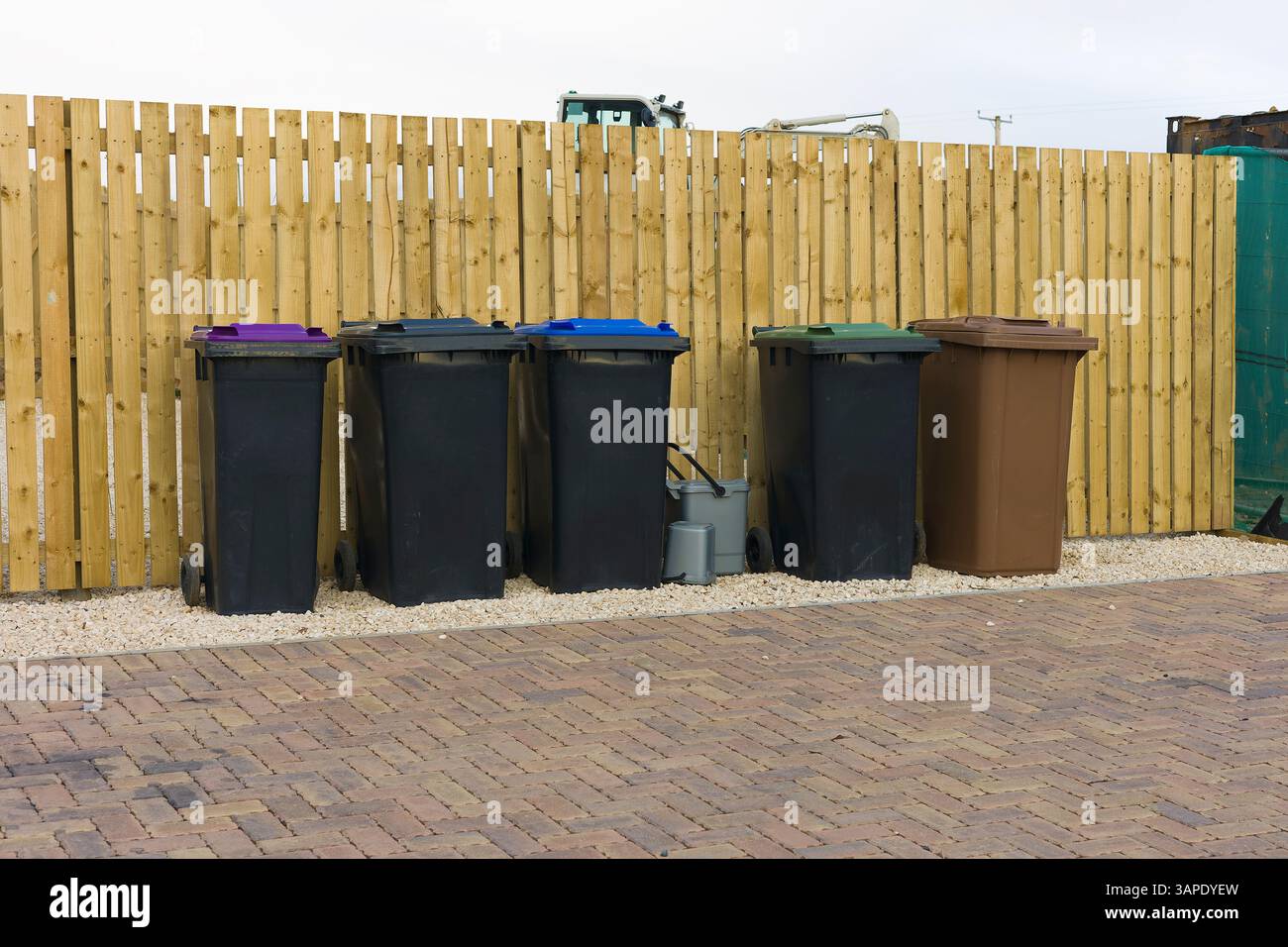 Fila di contenitori per il riciclaggio di rifiuti domestici in plastica con codice colore su lastre di pavimentazione di fronte alla nuova recinzione in legno del giardino nell'area residenziale Foto Stock