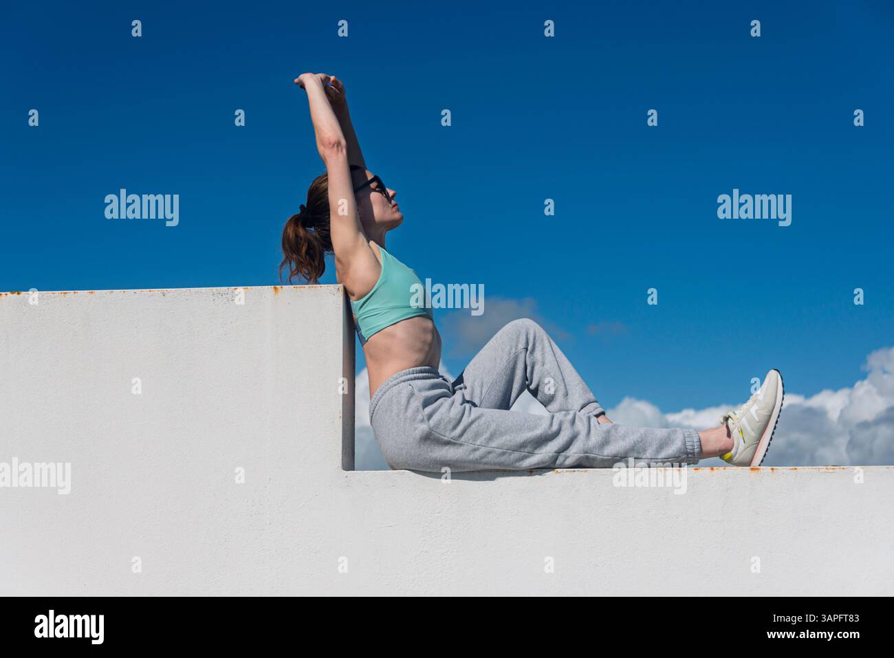 Cameriere sportivo seduto su una parete con le braccia sollevate che si allungano, si prende una pausa, Foto Stock