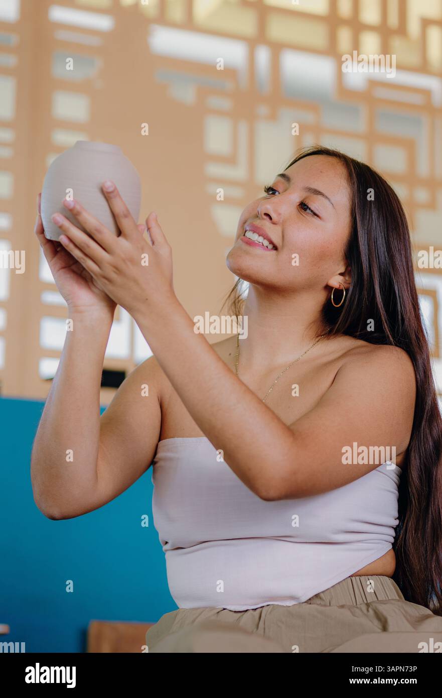 La giovane donna latina tiene e valuta i pezzi di ceramica che ha realizzato nel suo studio. Circondata da attrezzi, sorride calorosamente e indossa abiti semi-formali. Foto Stock