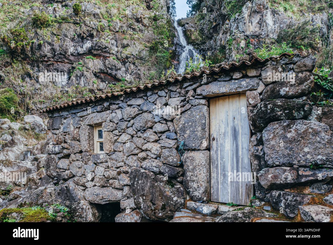 Un'affascinante casa in pietra in rovina si trova tra le rocce vicino a una splendida cascata a Sao Jorge, Azzorre, Portogallo. Una pittoresca scena di rus' Foto Stock