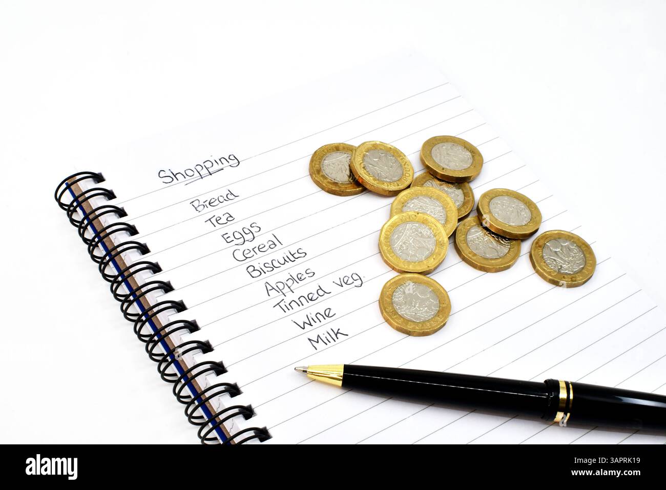 Vista ravvicinata di una lista della spesa scritta su una pagina di un notebool con carta foderata, con monete inglesi da una sterlina e una penna. Niente persone. Foto Stock