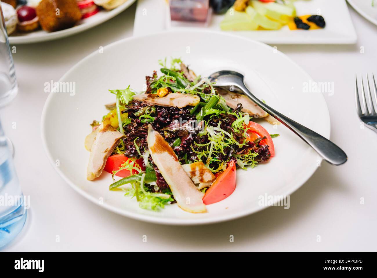 Insalata fresca di verdure a foglia verde, pomodori e fette di pollo alla griglia servita in un recipiente bianco con un cucchiaio su una tovaglia bianca. Foto Stock