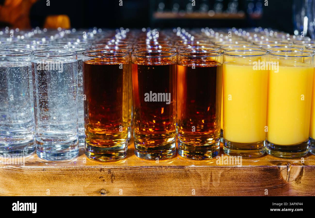 File di bicchieri pieni di acqua frizzante, soda scura e succo d'arancia allineati su un tavolo di legno per il servizio bevande. Foto Stock