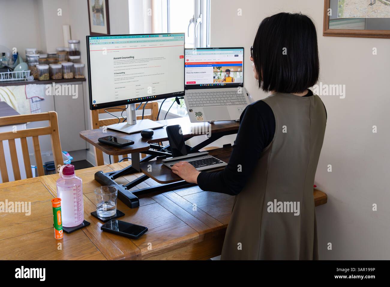 Donna in piedi a un tavolo da cucina con un convertitore da tavolo e con due schermi per computer, Regno Unito. Concetto: Equilibrio della vita lavorativa, lavoro flessibile, wfh Foto Stock