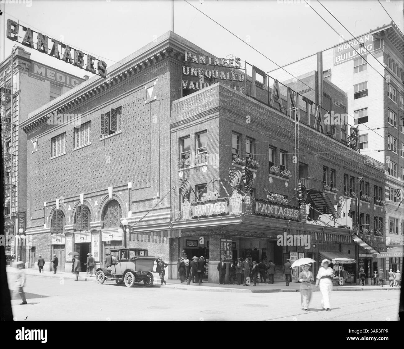 Questo ritratto all'aperto cattura la vita cittadina di Portland, Oregon, con un grande edificio in mattoni identificato come il Pantages Vaudeville Theater. La superficie del teatro è adornata da cartelli illuminati. Una folla si riunisce vicino all'ingresso mentre un uomo si appoggia su un'automobile parcheggiata. Due donne, una con un ombrello, attraversano la strada. L'edificio sulla destra è contrassegnato con "Morgan Building". Gli elementi aggiuntivi includono lampioni stradali, linee elettriche, fiori in vaso e una scala. Foto Stock