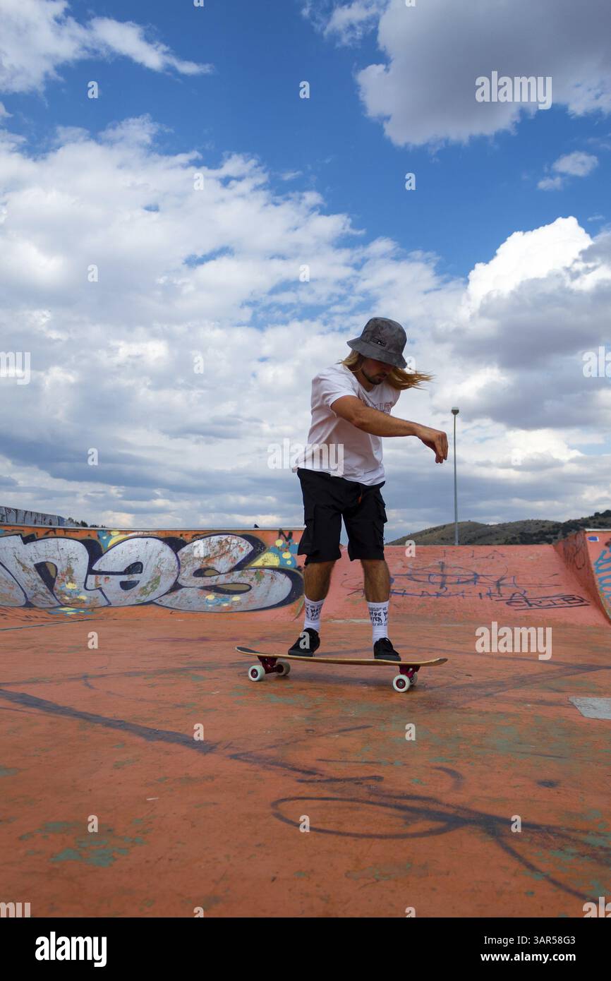 Uno skateboarder si concentra sul perfezionamento della sua tecnica in un vivace skatepark urbano, con uno sfondo di cieli blu e nuvole soffici Foto Stock