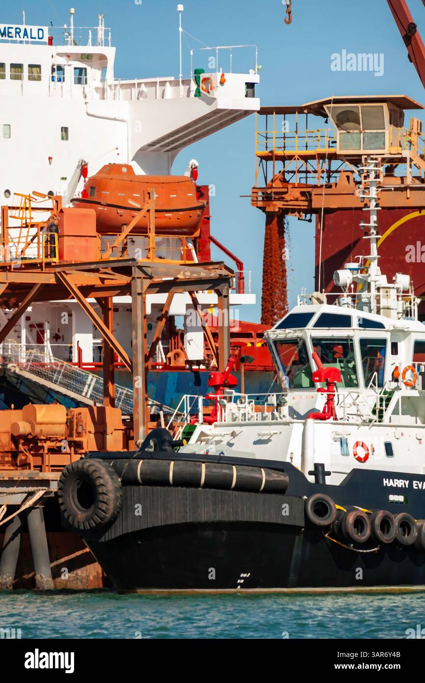 rimorchiatore ormeggiato da una nave color smeraldo presso un molo del porto con macchinari industriali attrezzature per lo scarico di materiali che esemplificano la vivace attività portuale Foto Stock