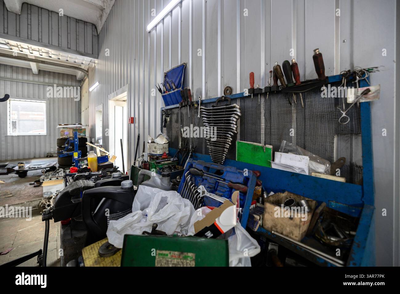 Un banco di lavoro ingombrante e un pannello di controllo con vari utensili e forniture per il settore automobilistico sono visibili all'interno di un'officina di riparazione molto affollata. Foto Stock