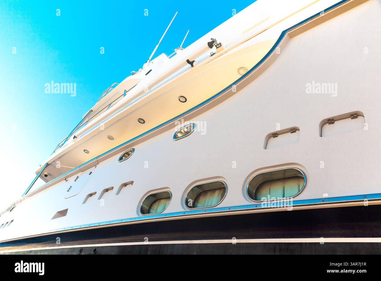 Lo scafo di un lussuoso yacht bianco a motore, con oblò sopra, contro il cielo blu e la luce del sole nell'angolo, vista dal basso. Foto Stock