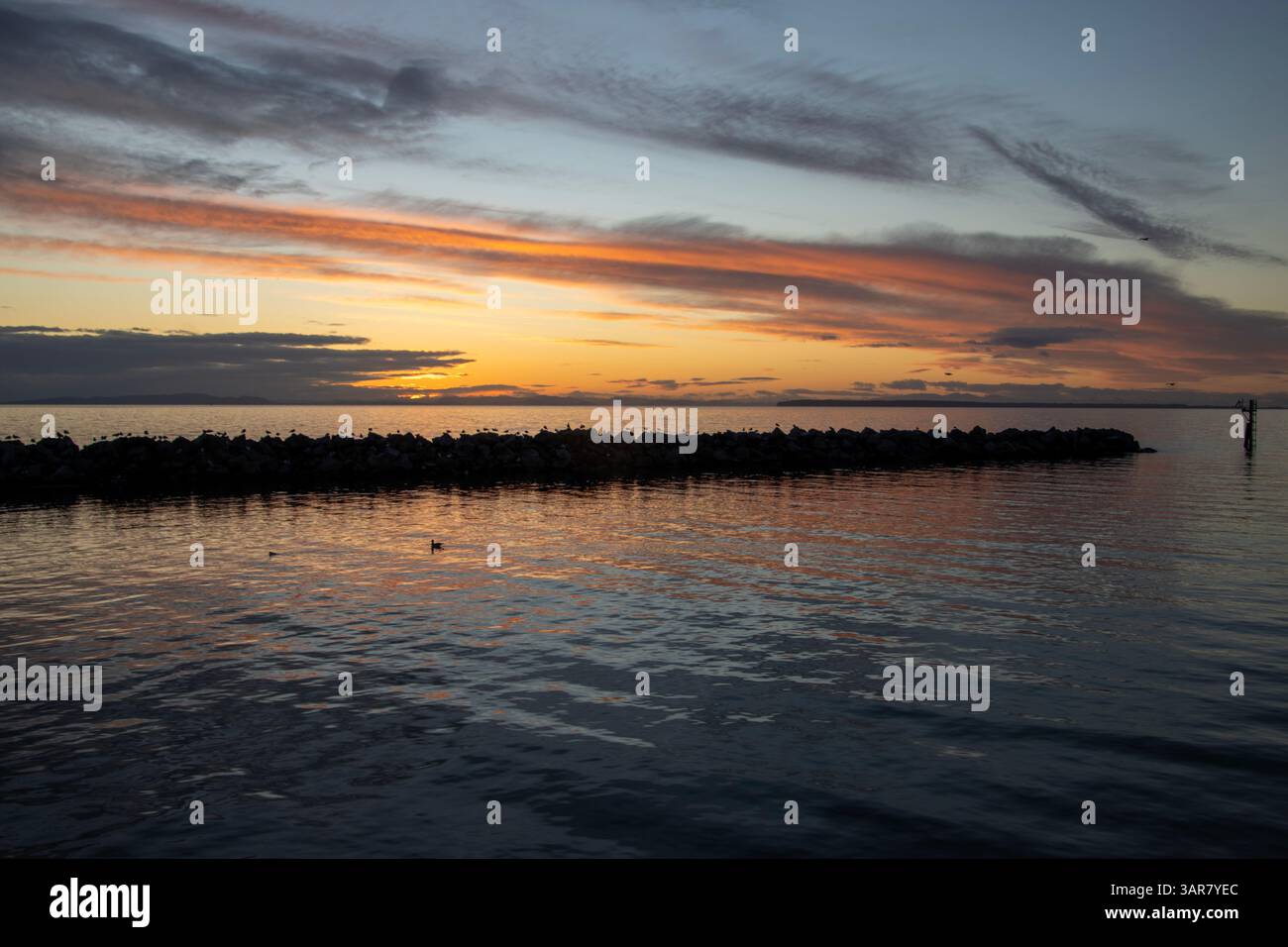 Un tramonto sereno si dispiega sulle acque tranquille, diffondendo vivaci riflessi di tonalità arancio e viola. Rocce sagomate e onde morbide valorizzano il pe Foto Stock