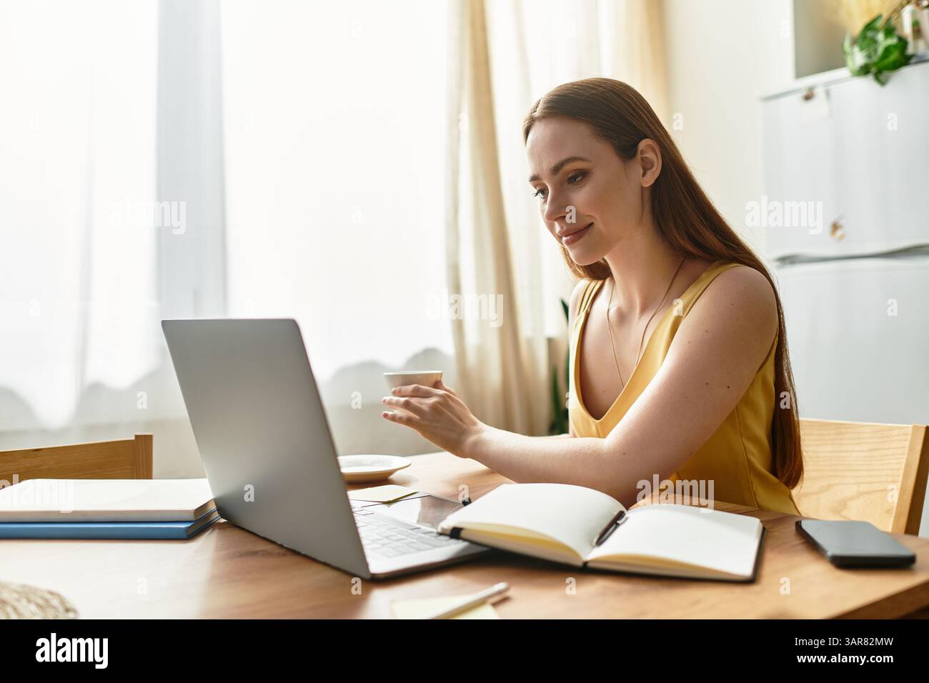 La giovane e bella donna sorride mentre partecipa a una riunione online presso il suo spazio di lavoro illuminato dal sole. Foto Stock