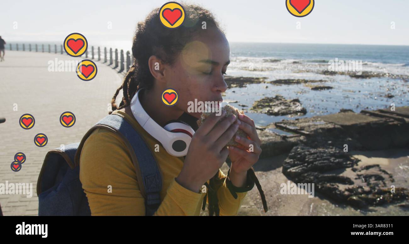 Molte icone del cuore fluttuano contro una donna afroamericana che mangia uno spuntino sul lungomare Foto Stock