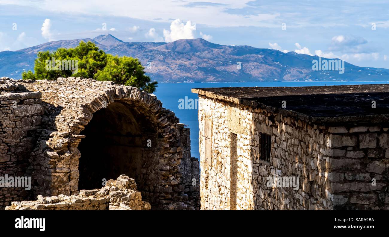 Una vista panoramica delle antiche rovine di pietra con uno sfondo di montagne e un mare calmo. Le rovine sono caratterizzate da una struttura ad arco e da un muro di pietra, circondato da un muro Foto Stock