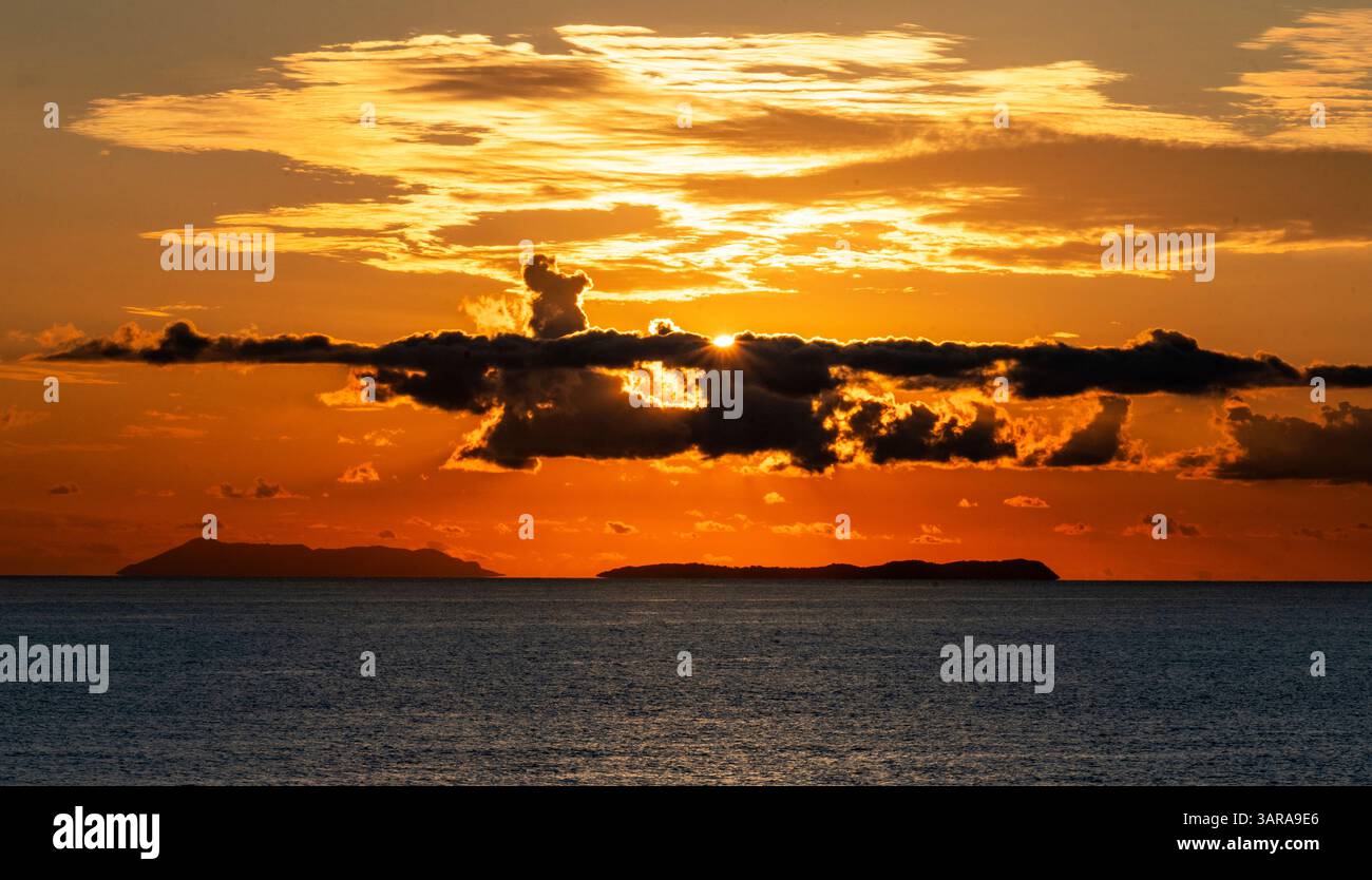 Uno splendido tramonto sull'oceano, con vivaci sfumature arancioni e gialle che illuminano il cielo. Le nuvole scure creano un contrasto drammatico, mentre l'isola è lontana Foto Stock