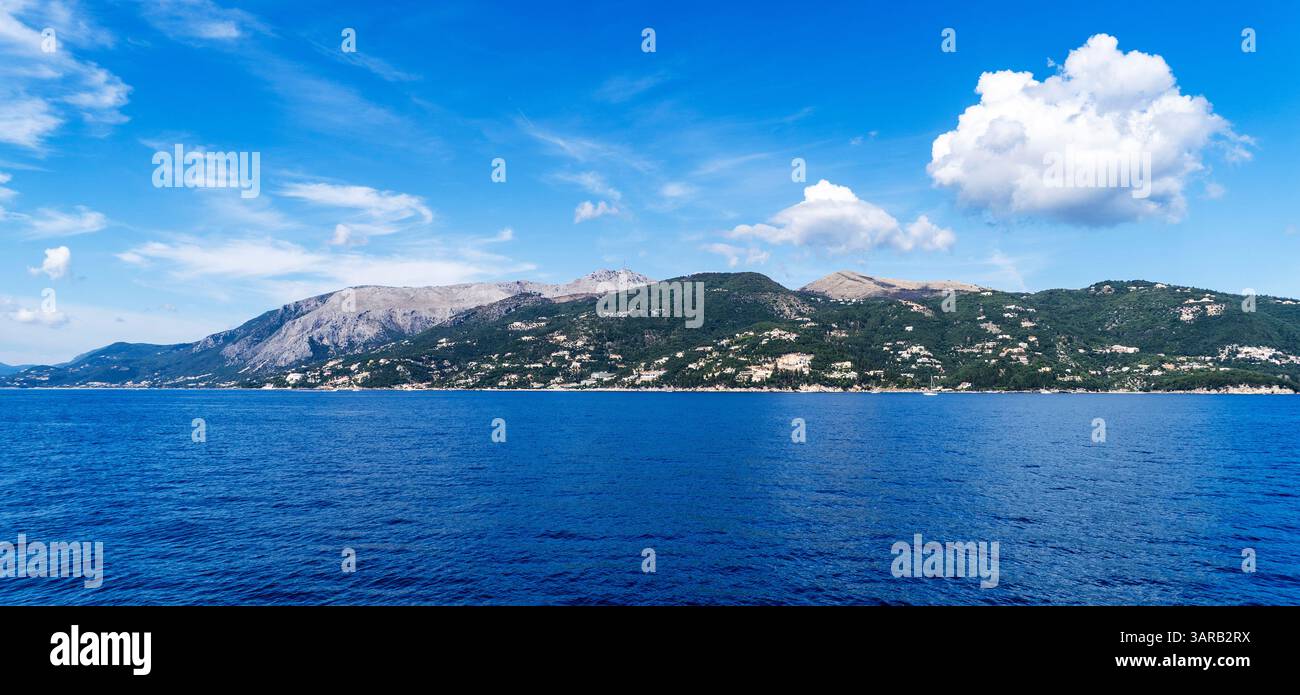 Una vista panoramica di una tranquilla costa con lussureggianti colline verdi e un cielo azzurro. Il mare calmo riflette i colori vibranti del paesaggio, creando Foto Stock