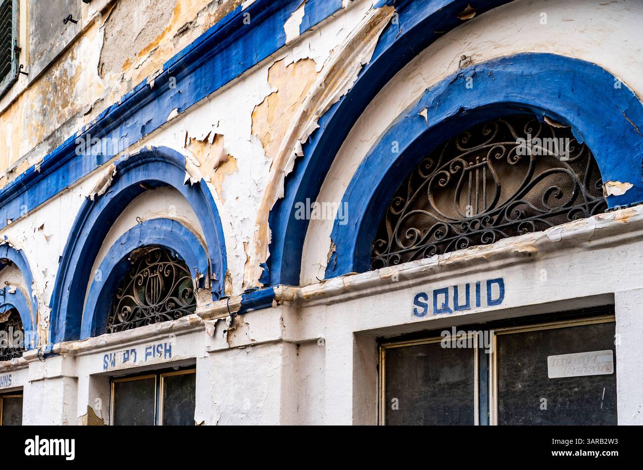 Una vista ravvicinata della facciata di un edificio intempestivo caratterizzato da finestre ad arco con lavori in ferro decorativi. La vernice si sta staccando, rivelando strati di blu e W. Foto Stock
